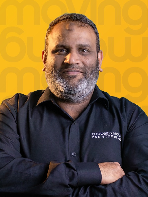 portrait of a man with a gray beard wearing a black shirt, arms crossed, against a yellow patterned background with a subtle logo motif.