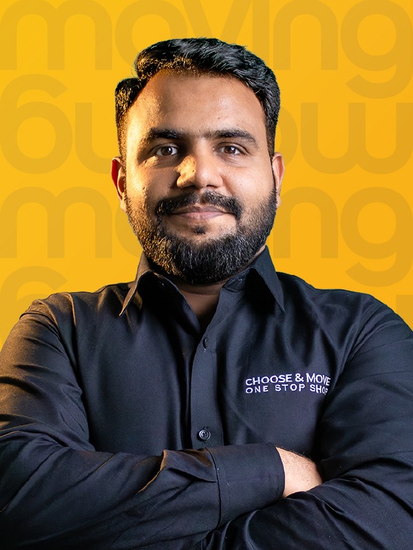 portrait of a man with short dark hair and beard, arms crossed, wearing a black shirt with 'choose & move one stop shop' logo, yellow patterned background