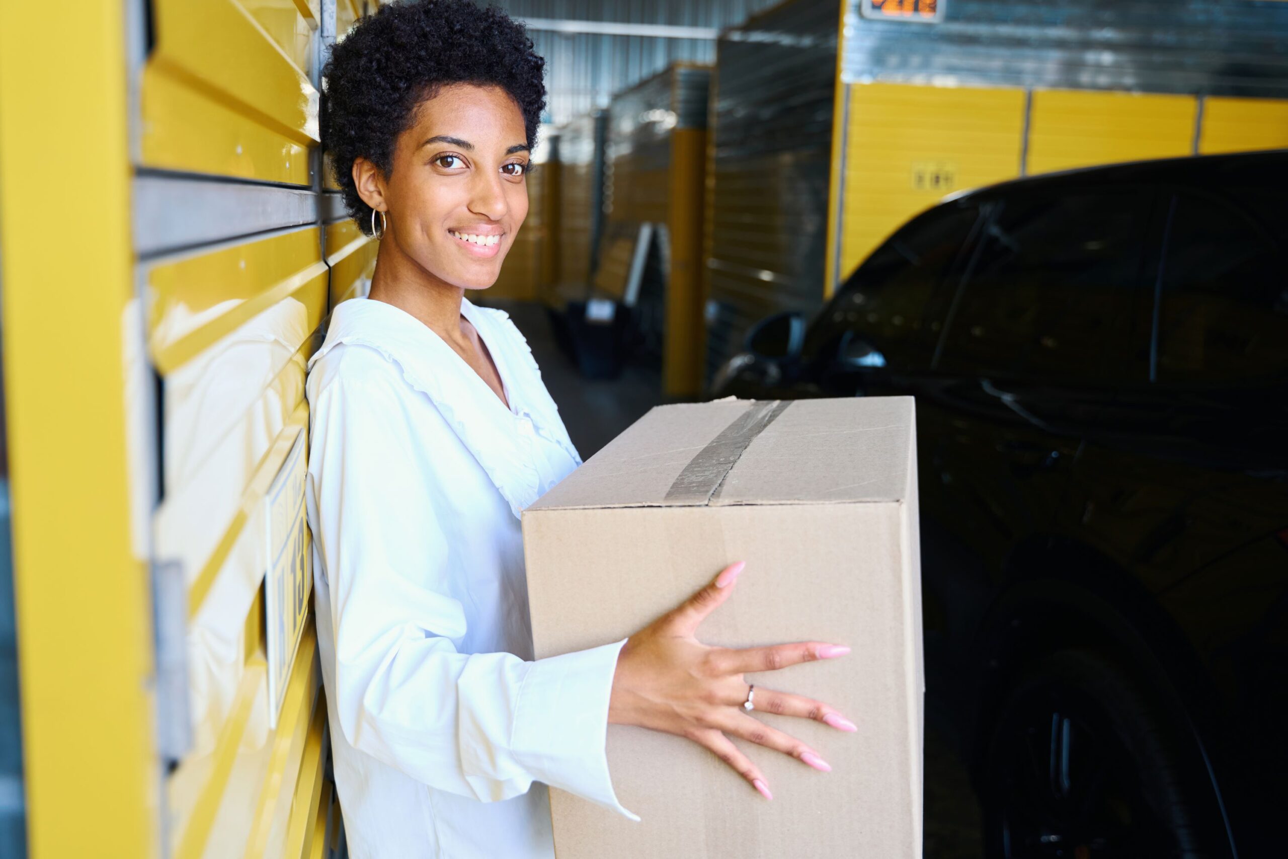 smiling woman in a white shirt leaning against yellow warehouse lockers while holding a cardboard box.