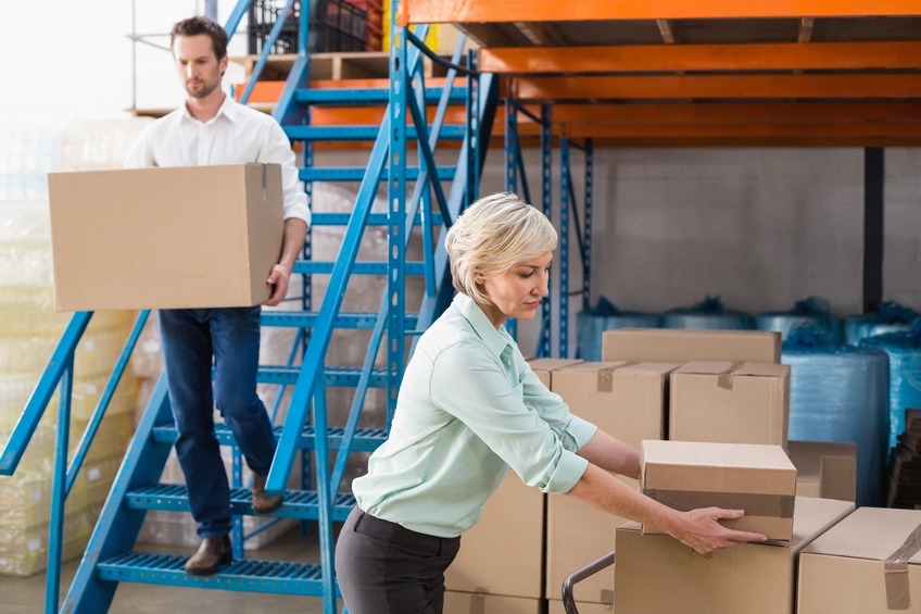 woman stacks cardboard boxes in a warehouse while a colleague carries a box down blue metal stairs in the background.