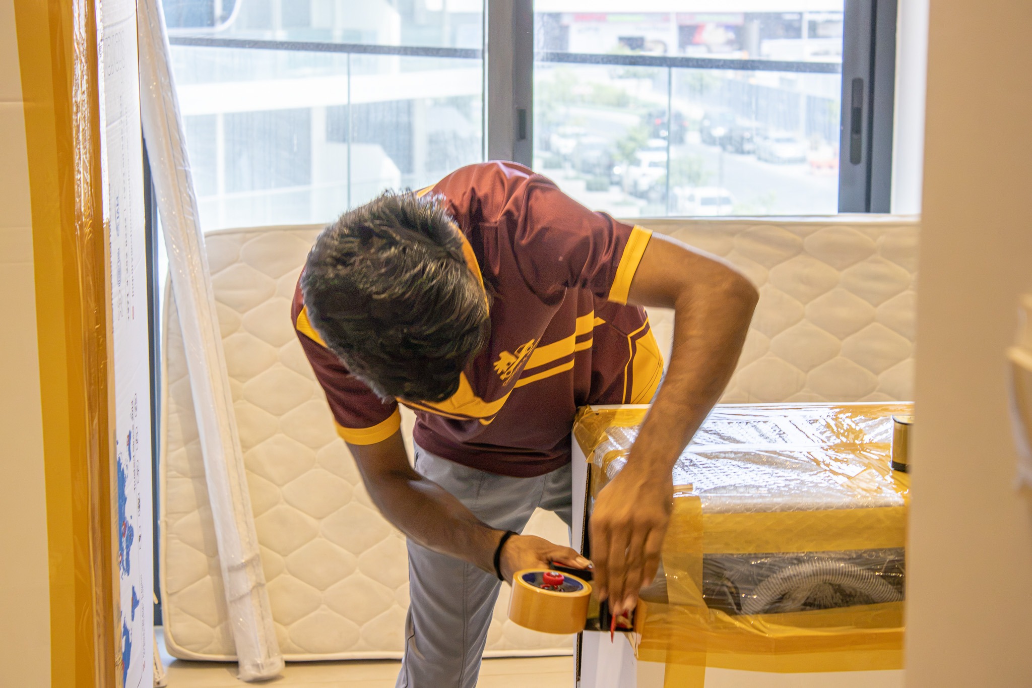 man in a maroon and yellow uniform seals a cardboard box with packing tape in a bright room with a mattress and window view behind him.