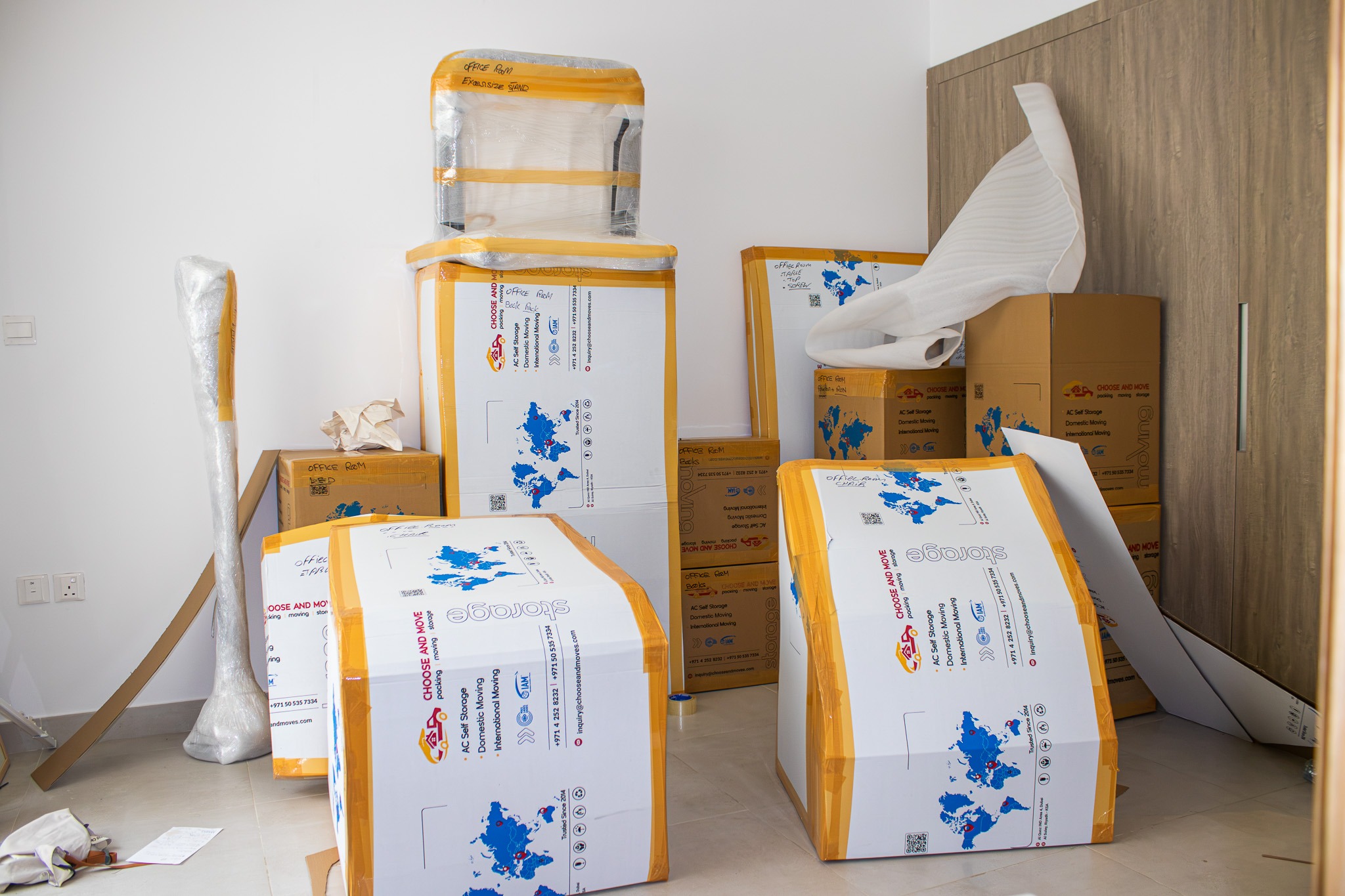 stacked moving boxes wrapped in orange tape against a white wall in a cluttered room, ready for transport.