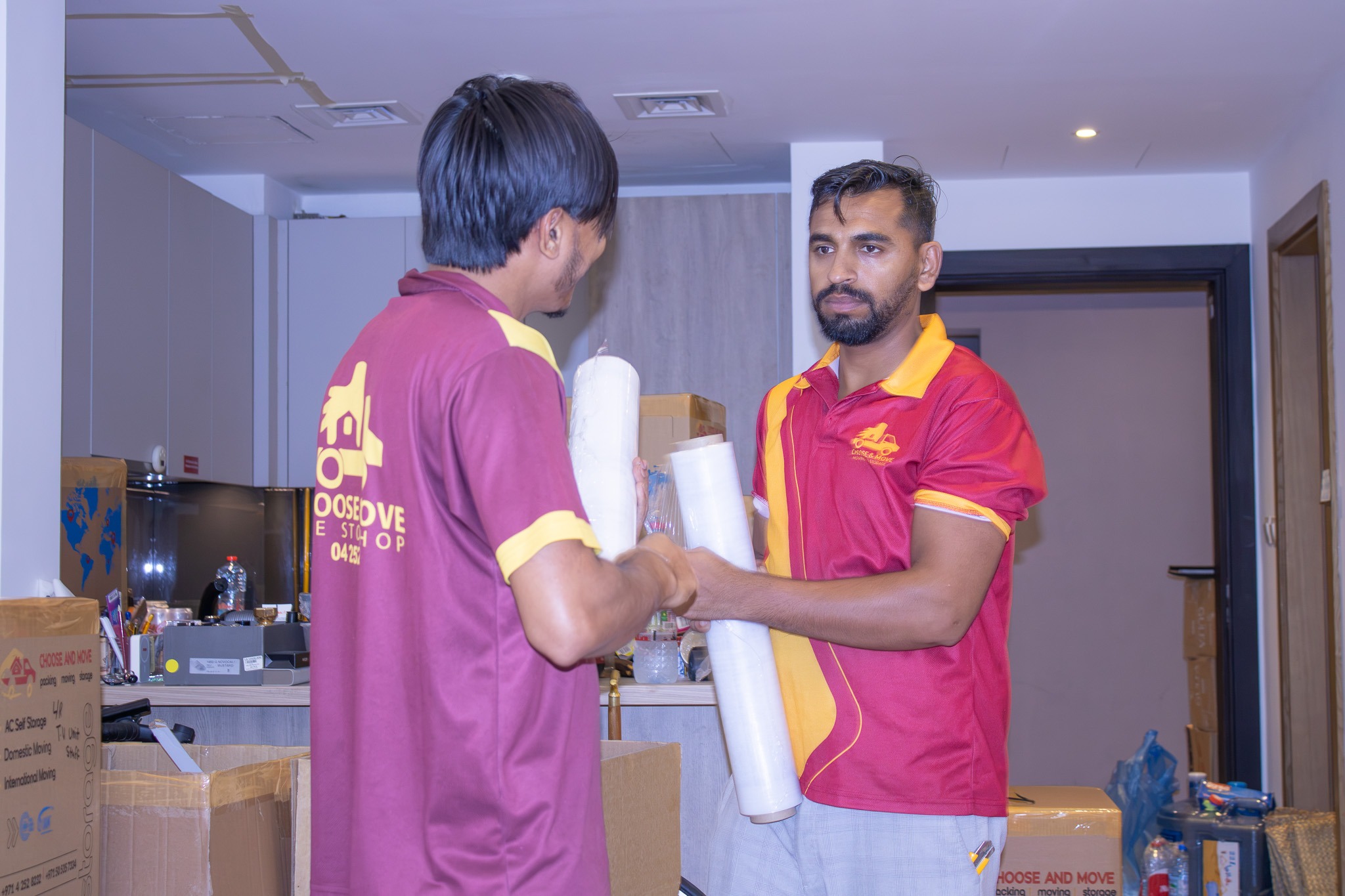two movers in maroon and yellow uniforms exchange packing materials in a cluttered packing room with boxes and supplies around them.