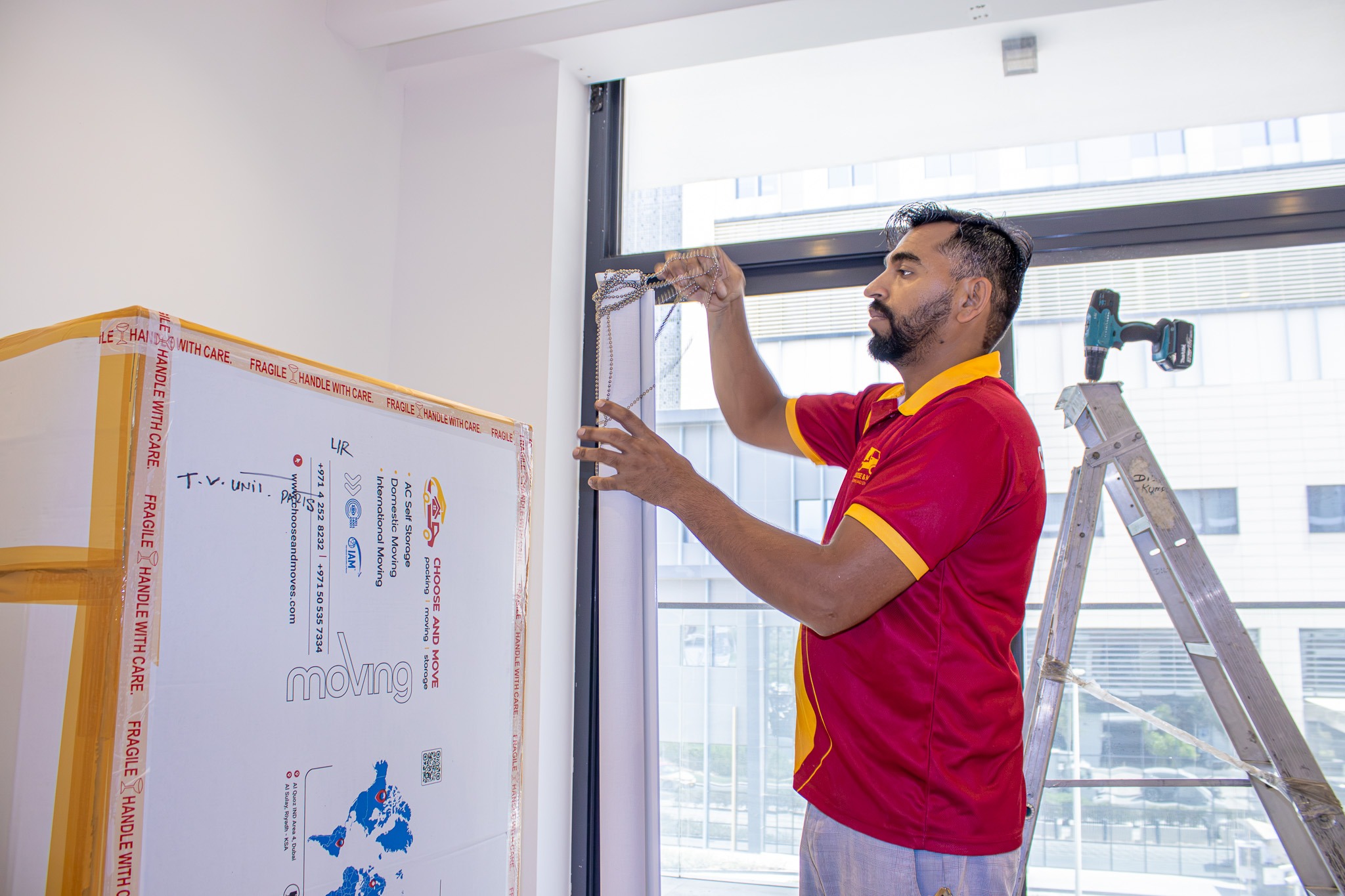 worker on a ladder installing a metal frame beside a large window, wearing a red polo shirt with yellow trim and gloves nearby a fragile-packed box.
