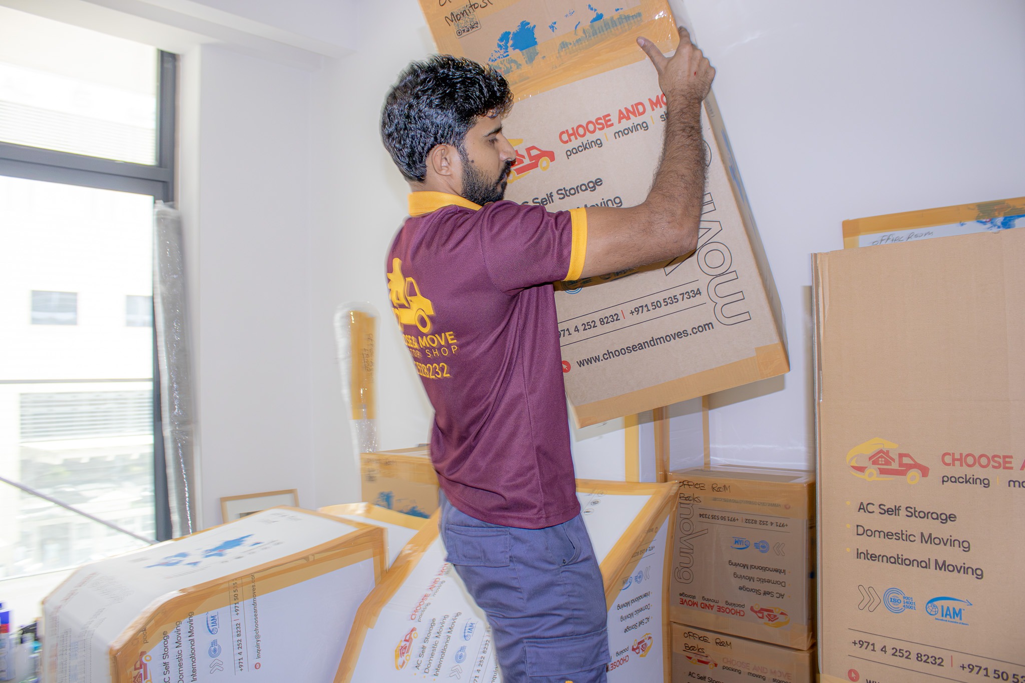 a man in a maroon shirt with yellow collar lifts large cardboard moving boxes in a room filled with stacked boxes for storage and packing