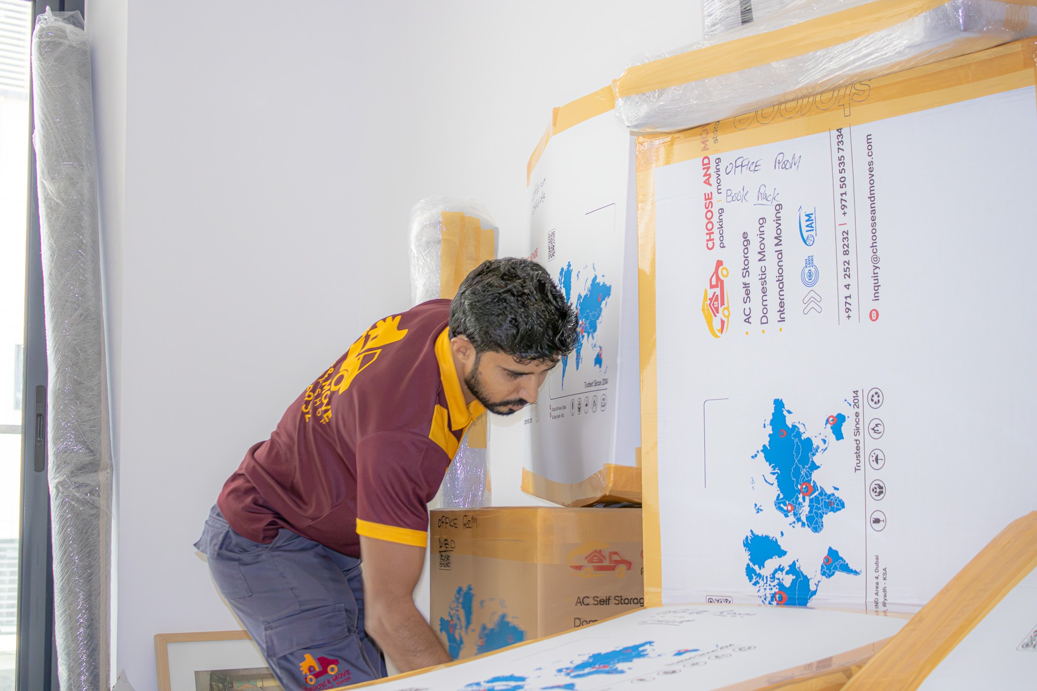 mover in a maroon and yellow uniform stacks large white moving boxes wrapped in tape in a bright room.