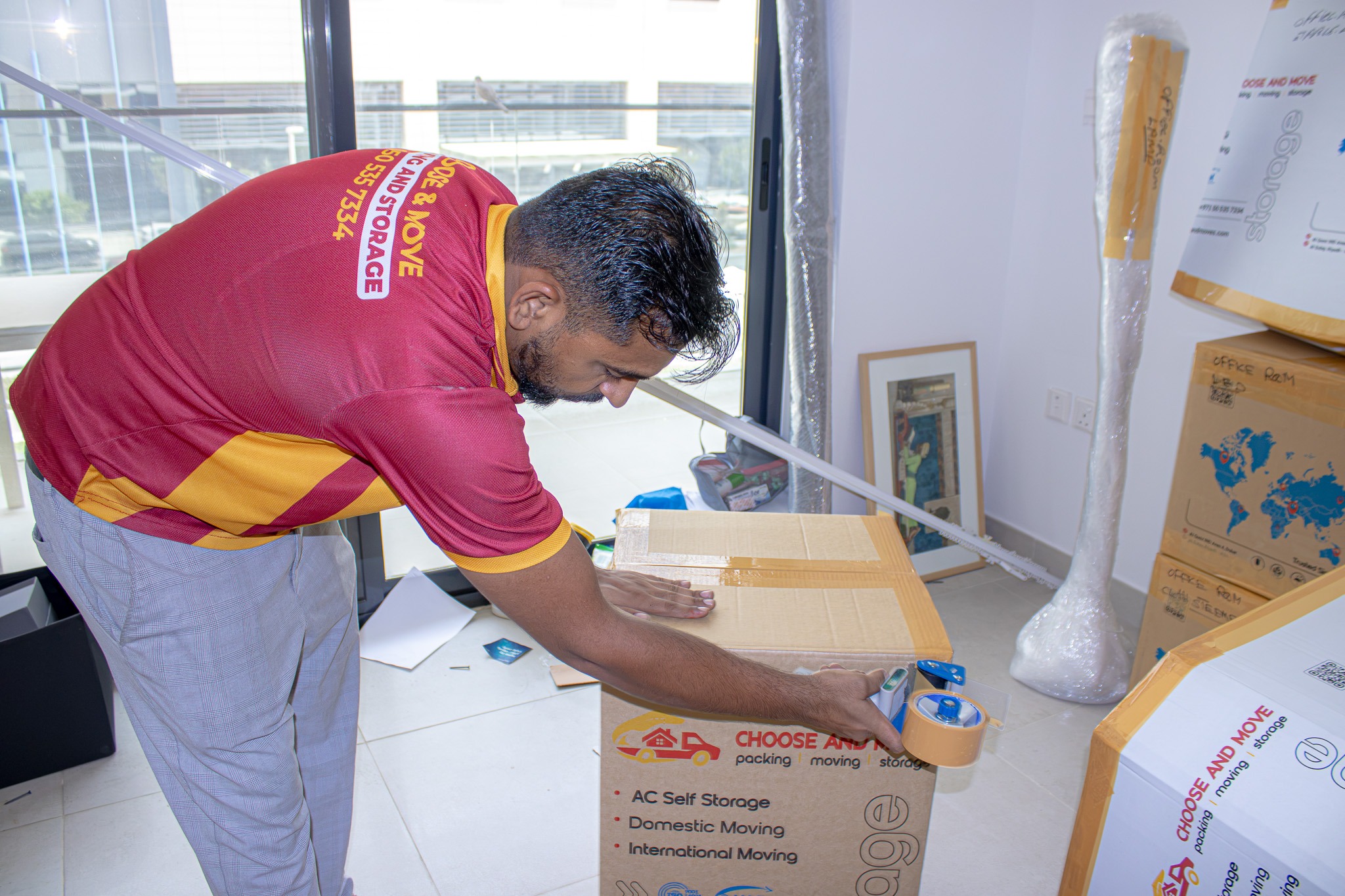 man in a red and yellow shirt seals a large cardboard box with packing tape in a bright storage room.