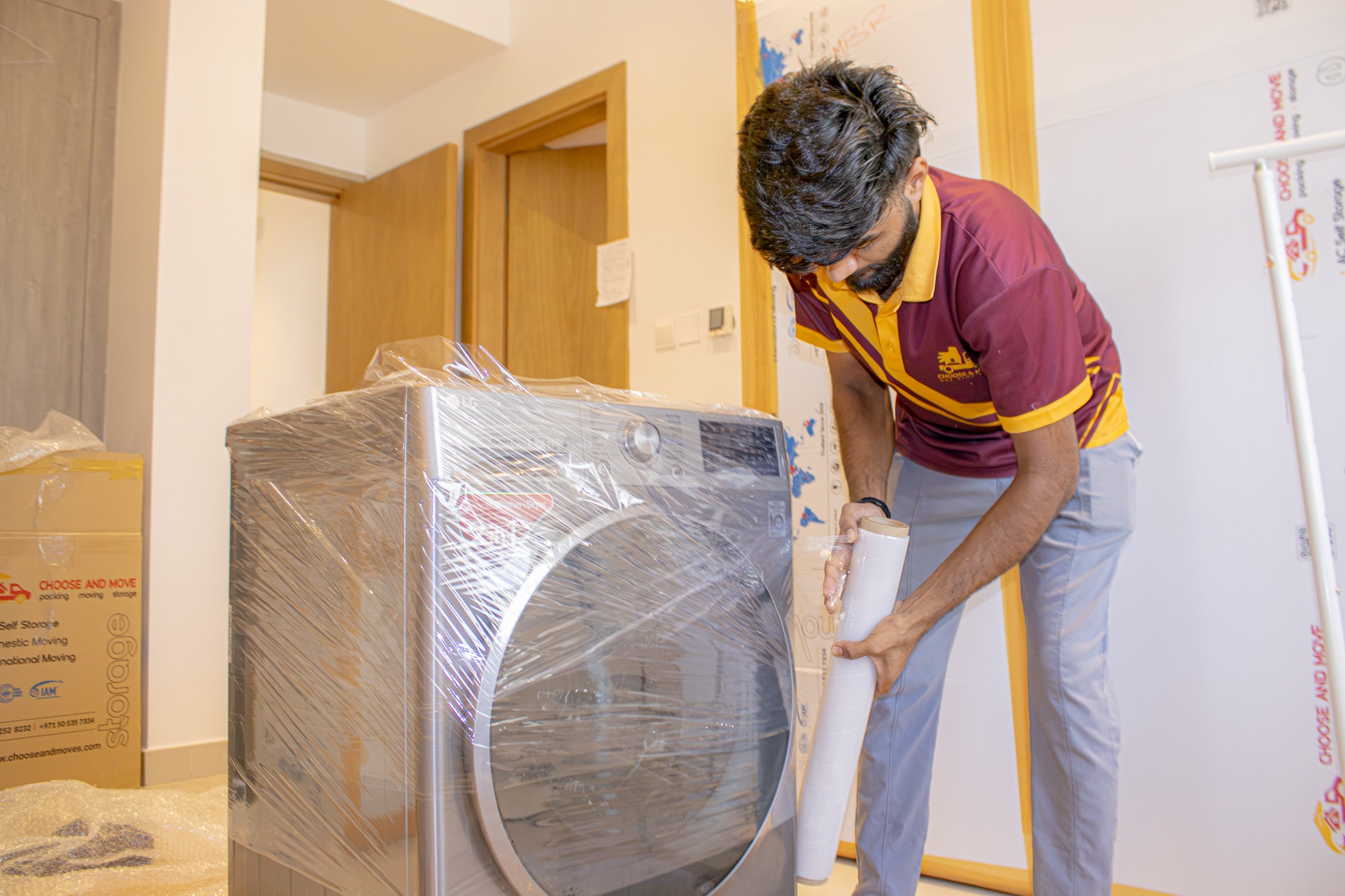 mover in maroon and yellow uniform inspecting a large appliance wrapped in plastic for transport in a storage room.